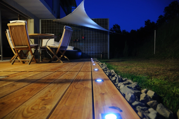 White waterproof triangular shade sail over a wooden garden deck with furniture at night.
