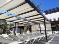 Copertină romană albă pe o pergolă închisă la culoare deasupra șezlongurilor de piscină într-o zi însorită