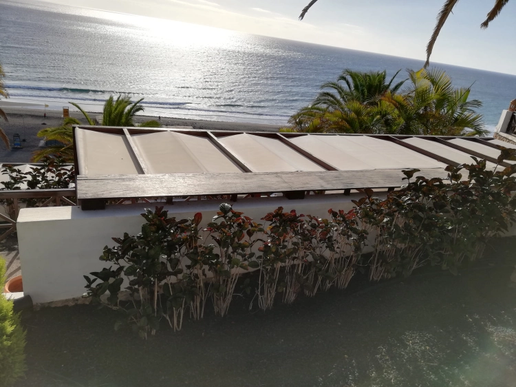Roman sun shade 2.1x7m installed over a terrace overlooking the beach and ocean with palm trees and green plants
