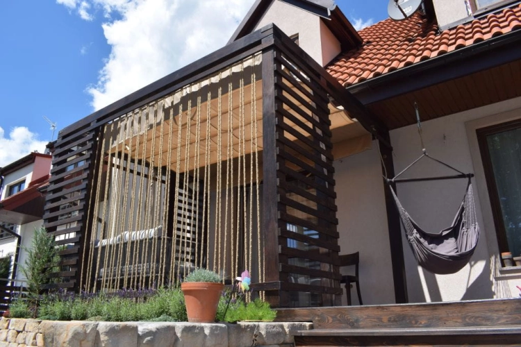 Outdoor beige Roman blind on a dark wooden terrace structure with a grey garden swing hanging nearby.