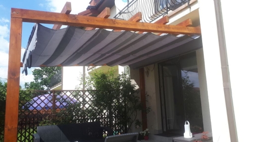 Grey and white striped retractable Roman shade canopy on a wooden pergola over a patio with garden furniture.