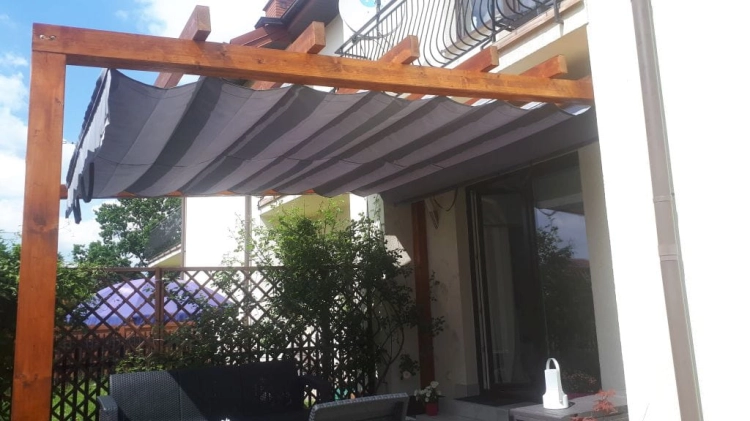Grey and white striped retractable Roman shade canopy on a wooden pergola over a patio with garden furniture.