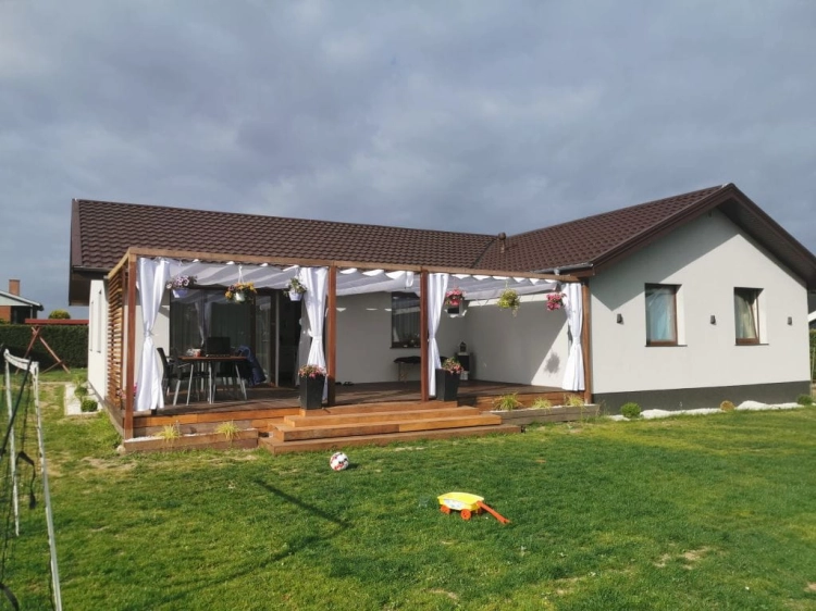White Roman outdoor shades installed on a wooden terrace pergola of a modern one-story house with a garden