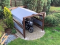 Graubeiges wasserdichtes Sonnensegel 2x2 m auf Holzkonstruktion über Motorrad auf Pflasterstein neben Pflanzen gespannt