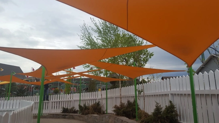 Orangefarbenes dreieckiges Sonnensegel Premium montiert über einem Spielplatz hinter einem weißen Zaun