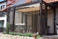 Estor romano claro para terraza de 4x4 m montado en una pérgola de madera con cuerdas decorativas y plantas en el jardín
