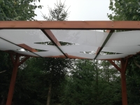 White sunshade sail strips woven through a wooden pergola structure against a backdrop of green forest trees.