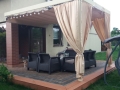 Cortina de jardín premium beige a medida colgada en una pérgola moderna con muebles de exterior