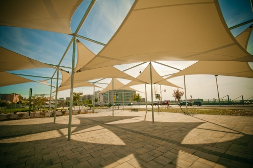 Large sand-colored triangular HDPE sun shade sails installed on a metal structure over a modern paved plaza area