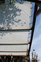 White custom-made rectangular sun shade sail mounted on a wooden pergola with shadow patterns of tree leaves.