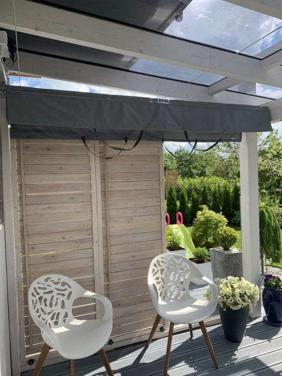 Grey horizontal retractable awning installed under a glass pergola roof on a wooden deck with white chairs.