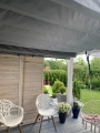 Retractable horizontal grey shade sail mounted under a pergola above a patio with chairs and garden view