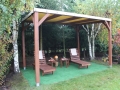 Fából készült pergola bézs PVC tetővel, két nyugággyal és asztalokkal a kertben a fűvön