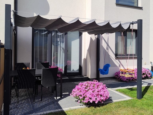 Grey 3x4 m premium waterproof wavy roman blind pergola with grey dining set, blue baby swing and pink flowers on a patio