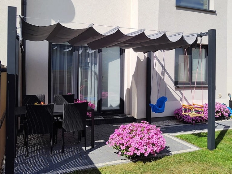 Grey 3x4 m premium waterproof wavy roman blind pergola with grey dining set, blue baby swing and pink flowers on a patio