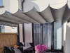 Retractable grey roman shade canopy over a patio dining set with a table, chairs, and pink flowers in the background