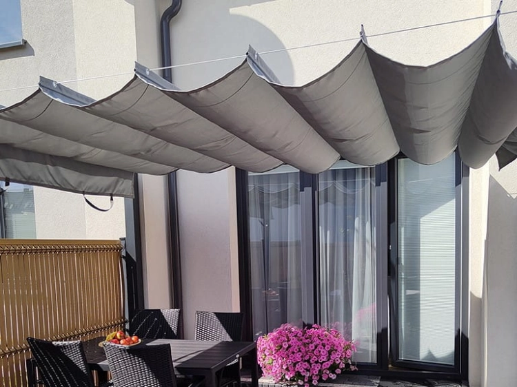 Retractable grey roman shade canopy over a patio dining set with a table, chairs, and pink flowers in the background