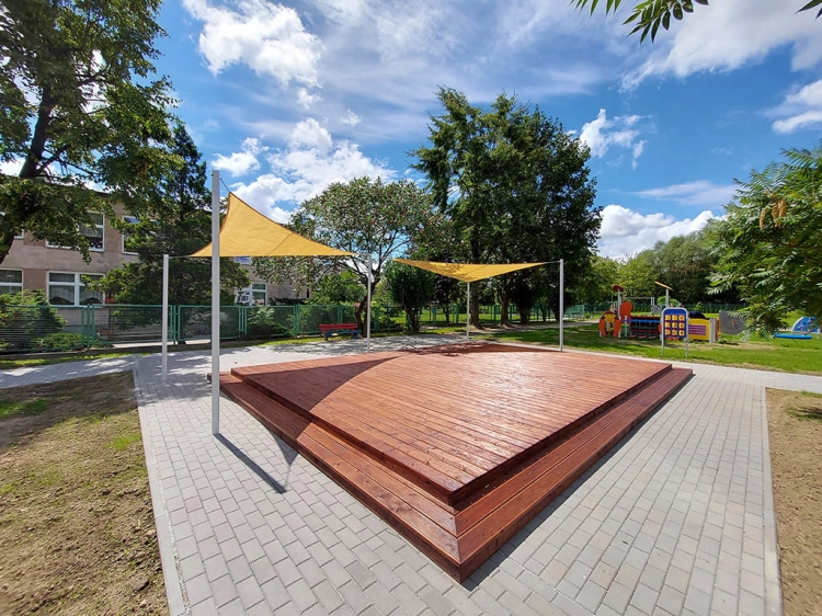 Yellow triangular sun shade sail installed over a wooden deck platform in a sunny outdoor park area.