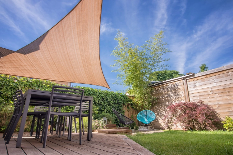 Triangular HDPE sun shade sail over a modern patio dining set in a lush garden with deck and lawn.