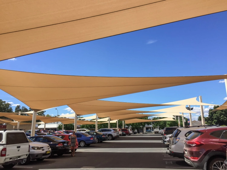 Beige triangular garden sun shade sail made of HDPE fabric installed over a parking area under a clear blue sky