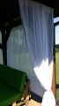 White sheer outdoor curtains for terrace or gazebo hung on a wooden beam near a green garden sofa.