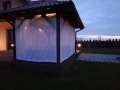 Wit gordijn voor terras dat een verlicht tuinhuisje afschermt in de schemering