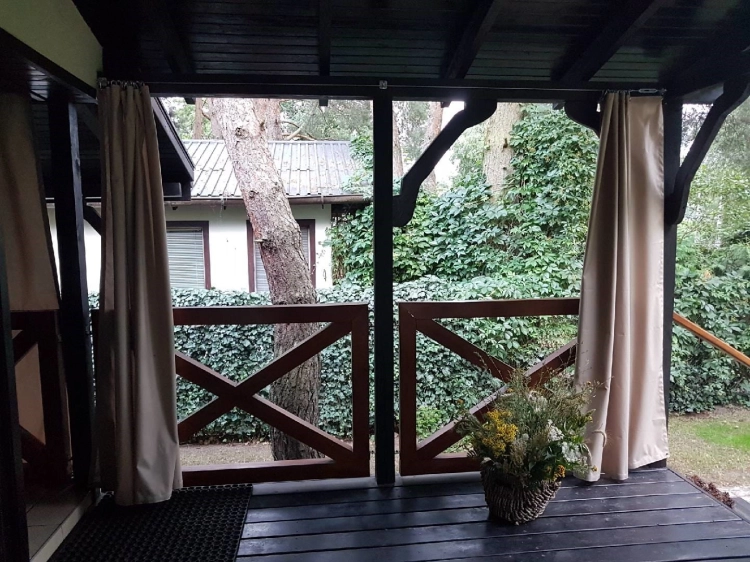 Beige waterproof garden curtains hung on a dark wooden terrace with a view of greenery and a house.