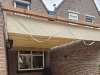 Cream waterproof retractable Roman shade for terrace installed over a patio between brick buildings.