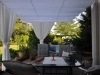 White Roman blind patio canopy with side curtains over a furnished terrace with a dining table and garden greenery