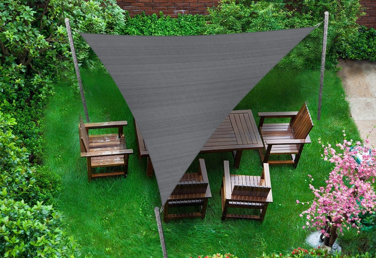 Triangular grey sun shade sail over a wooden garden furniture set on a green lawn with trees and pink flowers.