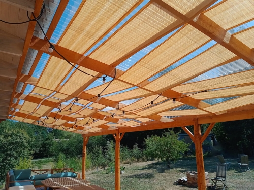 Beige HDPE sunshade fabric strip woven through a wooden pergola structure over a patio with outdoor furniture