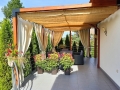 Customized HDPE Roman blind for a terrace pergola providing shade for outdoor furniture and potted plants.