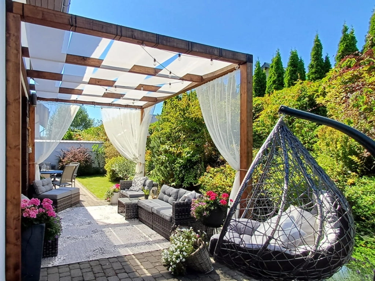 Custom-sized white sunshade fabric strips woven through a wooden pergola with outdoor lounge furniture and curtains.