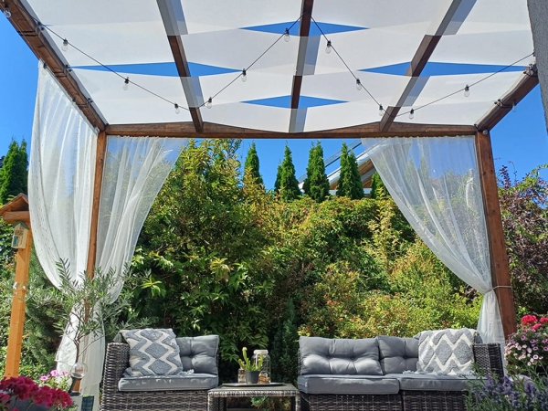 Custom white sunshade sails woven through a wooden pergola over a garden lounge set with grey cushions and white curtains