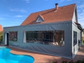 Grijs, transparant zeil op maat ter bescherming van het terras bij het zwembad en het huis met pannendak