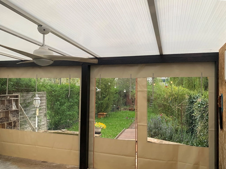 Priehľadná plachta na terasu s béžovým okrajom a oknom, namontovaná pod zastrešením s ventilátorom