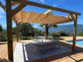 Tan retractable sunshade wave sail mounted on a wooden pergola over a poolside patio with outdoor furniture.