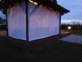Custom-made white sheer outdoor curtains for a wooden garden gazebo overlooking a yard at dusk