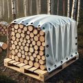 Custom-fit white tarp covering a large stack of firewood on a wooden pallet in a forest setting