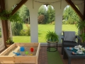 Transparent tarp with a white arch print covering a porch with a sandbox, outdoor furniture, and garden view