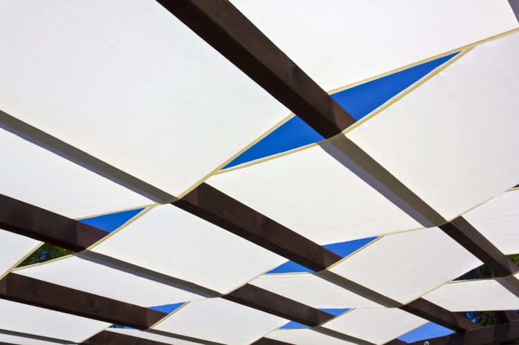 White waterproof sunshade sails woven through a dark wooden pergola frame against a clear blue sky