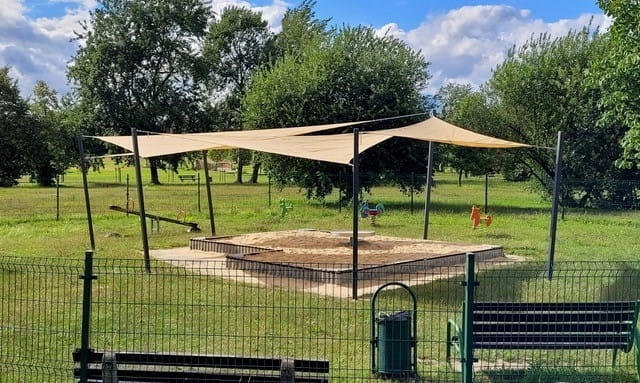 Tan sun shades covering a sandpit at a fenced playground with grass and trees in the background.