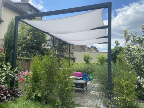 White waterproof sun sail canopy attached to a dark grey metal pergola over a garden patio with green plants.
