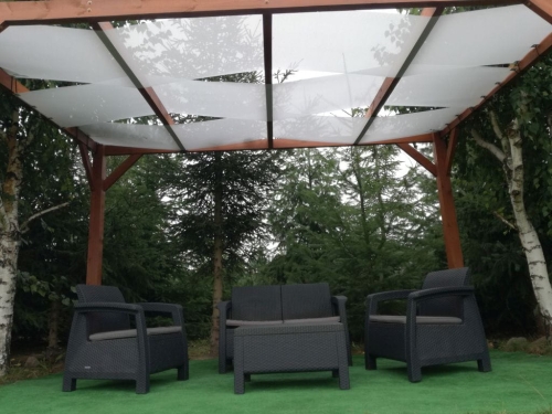 White outdoor sunshade fabric woven through a wooden pergola above dark wicker patio furniture on green grass