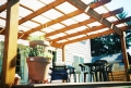 Beige outdoor sunshade sail strip installed over a wooden pergola on a backyard patio with a grill and dining set