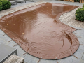 Custom-shaped brown winter pool cover secured with bungee cords on a stone and wooden deck patio