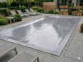 Custom rectangular gray vinyl pool cover secured with bungee cords on a stone patio with lounge chairs.
