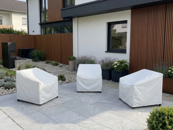 Three custom white waterproof covers protecting modern patio armchairs on a stone terrace in a landscaped garden