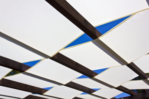 White waterproof pergola shade sail strips woven through dark wooden beams against a clear blue sky.