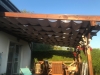 Beige premium waterproof Roman blind installed under a wooden pergola on a sunny backyard terrace holding outdoor furniture.